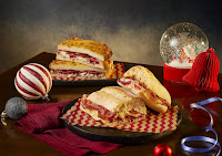 A wide angled photo of a selection of sandwiches with golden brown bread and various coloured meats and sauces on small white circular plates on a dark brown rectangular table on a bright background