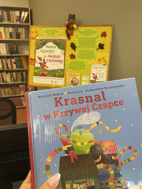 Mała książka - wielki człowiek_Wyprawka Czytelnicza Na pierwszym planie książka "Krasnal w krzywej czapce" Wojciecha Widłaka (na niebieskim tle widoczne cztery domki, na jednym z nich siedzi krasnal w czapce i długim pasiastym szaliku). Dalej widać tablicę informującą o projekcie Mała książka - wielki człowiek. Obok tablicy widoczny fragment regału z książkami.