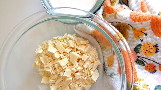 a glass bowl full of chopped white chocolate next to a dish of cream and a kitchen towel