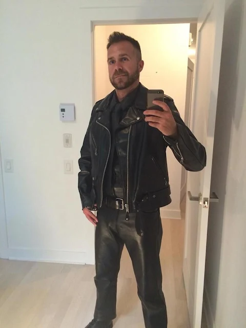 Taking a selfie in a white room with nothing in it a man wearing black leather pants and jacket smiling
