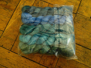 A clear plastic bag containing 5 small skeins of yarn, ranging from dark blue to green in a gradient.