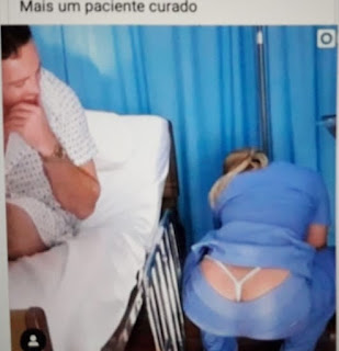 Foto. Em um box de enfermaria com divisória cortinada azul, à esquerda o paciente de camisolão claro com estampas pequenas acinzentadas e relógio dourado no pulso esquerdo, leva a mão ao queixo encobrindo o discreto sorriso. Ele observa à direita a enfermeira que está de costas. Agachada, ela ajeita o tripé do soro ao lado da maca, o jaleco azul sobe até a cintura e a calça de cós baixo deixa à mostra o avantajado bumbum demarcado pelas tiras do fio dental branco. No topo lê-se: Mais um paciente curado.