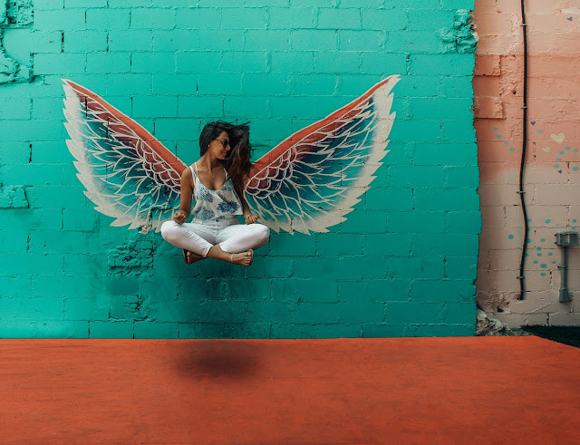 Woman levitating in mid-air, surrounded by a halo of light, with a mural of an angel on the wall behind her