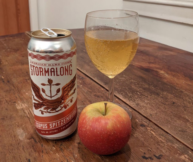 A wineglass of orange-gold cider with the can it came in and the apple it was made from