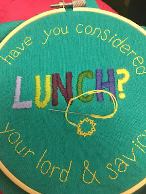 A plain wooden embroidery hoop with a dull silver screw, holding turquoise fabric embroidered with "Have you considered your lord and savior" in small yellow backstitch around the edges of the circle, and "lunch" in large satin-stitch capital letters of lavender, two shades of green, garnet, and navy. "Lunch" ends in a bright-yellow question mark. Two rows of bright yellow petals for a small sunflower are just off-center below "lunch," with a threaded needle set on top of the work.