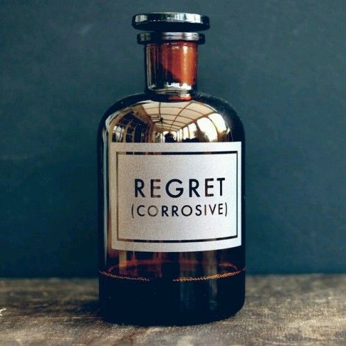 A brown glass apothecary bottle with a white etched on label that says Regret, (corrosive). There is a reflection of a bright glass conservatory roof which gives it a lightness. It is on a shelf with a blue wall behind. Take by Andy Poplar who makes them for his company Vinegar & Brown Paper.