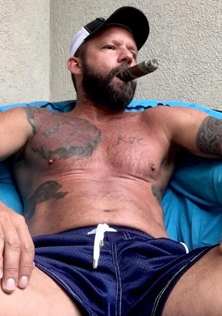 Tattooed middle-aged man wearing blue swimming trunks shirtless smoking a cigar sitting down