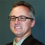 In three quarter profile, Mr. Miller poses with a smile in a black suit and green shirt and reading glasses on his face.