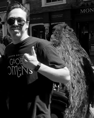 Black and white photo of a man whearing sunglasses, a black T short reading 'good omens' and black wings 