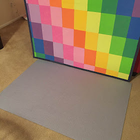 Large puzzle laying face down on a carpeted floor with a rainbow colored framed puzzle mat standing behind it