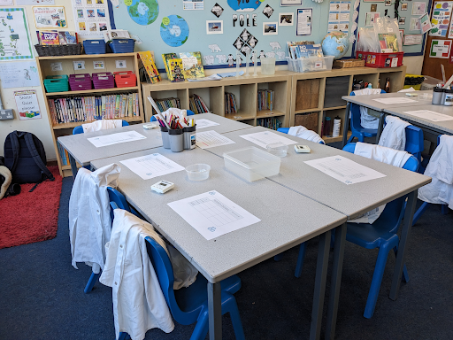 A photograph of a table in an empty classroom. There are chairs around the table and papers and writing equipment on the table