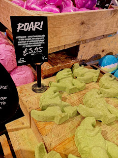 A pile of light green dinosaur shaped bath bombs on a square light brown box with a black rectangular table with roar! in white font on a bright background 