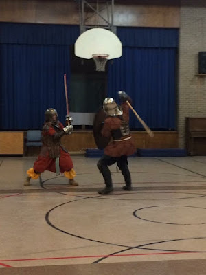 Two people in medieval garb and armour, in a school gym. The larger one in brown and black is holding a shield and ready to swing a sword over their shoulder at the smaller one in red and yellow, who's set to parry with a sword.