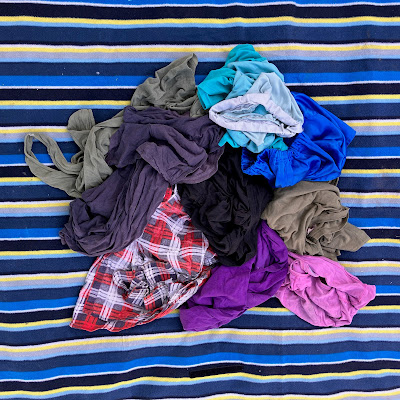Clothes that need mending have been tipped into a rough pile on a striped blue picnic rug. There is an olive tank top, a navy t-shirt, a child's ombre blue skirt, a child's mid-blue satin-look skirt, a plaid long-sleeved shirt in red, blue, and white, a black t-shirt, a purple t-shirt, an olive t-shirt, and a child's pink long sleeved t-shirt.