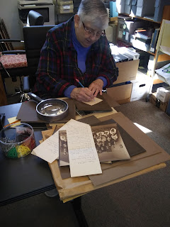 An older woman sits at a desk writing notes about the historic photos spread out in front of her. A magnifying glass sits on the desk to her right.