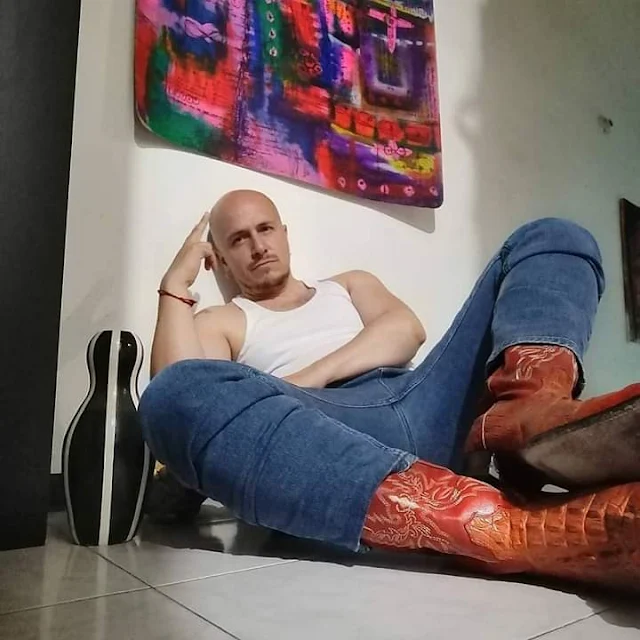 6/11 Bald thug sitting in bedroom on floor wearing blue jeans, a white wife beater and orange cowboy boots with his hand on his head thinking seriously