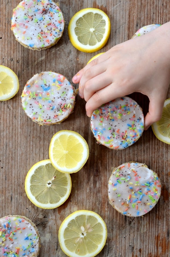 Runde Kuchenstücke mit Zitronenglasur und Zuckerstreuseln auf einem Holzbrett. Dazwischen liegen Zitronenscheiben. Eine Kinderhand greift nach einem Kuchenstück.