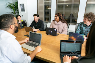 CC-BY-Mapbox-Uncharted-ERG_Mapbox-b024. Group of people in business casual, with their laptops, around a conference table. Group includes mix of genders, races and LGBTQ people.