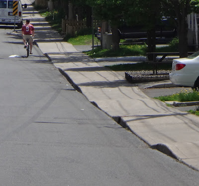 Looking along a sidewalk with the curb edge silhouetted showing frequent dips and bumps along the length of the sidewalk. (LeBreton Street North between Willow Street and Empress Street, looking north on the east side)