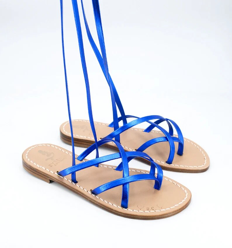 Low lace-up gladiator sandals Syrenia Sorrento, with cross on the instep, in blue laminated leather and leather sole. The laces are held by someone not present in the photo, towards the top, while the sandals are on a white background.