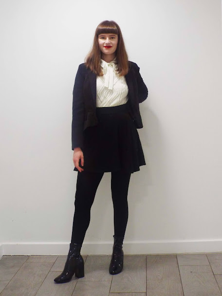 Ellie posed in black, on trend colour for AW20/21, skirt suit, paired with white pussy bow shirt and jumper, thick black tights and chucky, high heel, black PVC boots. Wearing hair down in a long bob, full fringe and a red lip.
