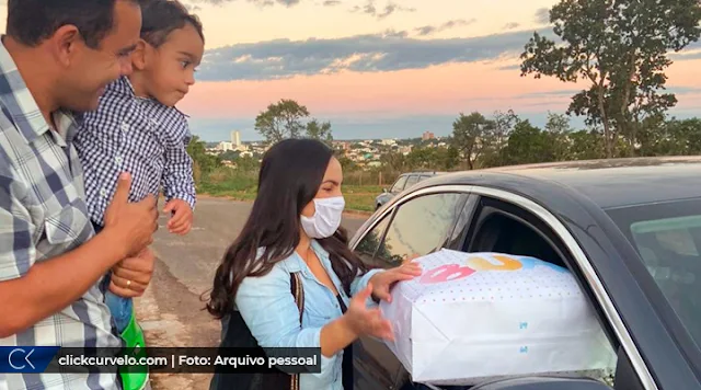 O pai Henrique Martins com o pequeno Henrique no colo ao lado da mãe, Karla Araújo, que recolhe um pacote de um carro. Todos sorrindo.