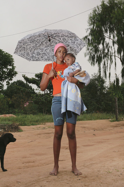 Uma mulher negra de pele clara é vista em pé com um bebê ao colo, olhando diretamente para a câmera com uma expressão séria. Ambos estão iluminados por flash direto. A mulher está descalça e traja shorts, uma camiseta laranja e uma touca rosa na cabeça. Segura o bebê enrolado em uma manta com o braço esquerdo e um guarda-sol com o braço direito. O chão é de terra; há um cão preto parcialmente ao lado esquerdo da imagem e  há vegetação e cabos de energia elétrica ao fundo. 