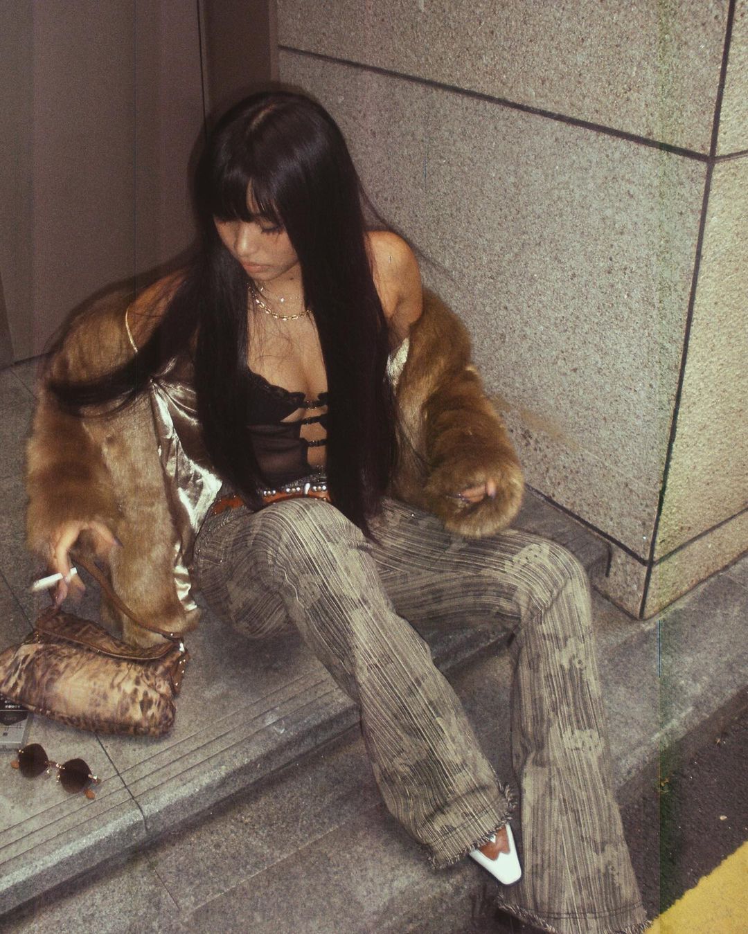 Woman with long straight black hair smoking a cigarette and wearing a brown faux fur jacket, grey flared jeans, leopard print baguette bag, and cowboy boots