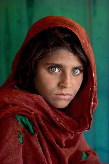 A imagem mostra o rosto de uma jovem de origem afegã, com um tecido de cor terrosa  enrolando sua cabeça, e seus olhos verdes muito vívidos e penetrantes olhando diretamente para a câmera fotográfica. O fundo do retrato se encontra desfocado, mas parece ser uma parede de madeira pintada na cor verde escuro. 