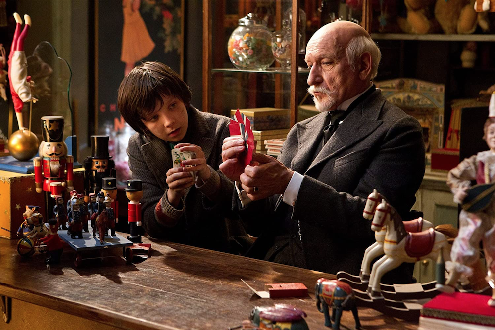 Asa Butterfield as Hugo Cabret and Ben Kingsley as Georges Méliès at counter in toy shop adorned with nutcracker soldiers, miniature rocking horses, and other hand-crafted figurines