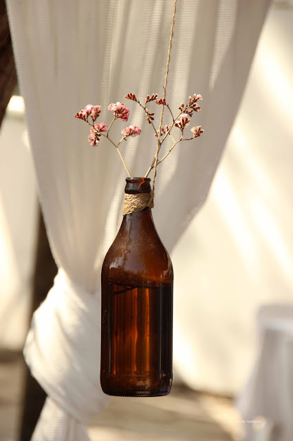 A brown glass bottle decorated with pink dried flowers hangs from a string of parcel ribbon, in front of white fabric.