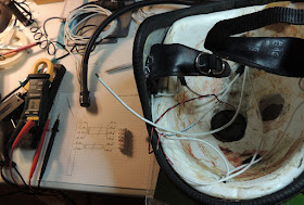 The helmet is upside-down on a cluttered workbench. The Styrofoam crush layer inside has wires running from place to place, powering the LEDs. The bench has a coil of more wire, a multimeter, a glue gun, pen and paper with a terminal diagram, and a screw terminal block.