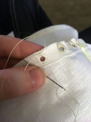 A close-up of a white finger and thumb supporting the narrow double-folded hem of a piece of white linen, with a needle pinched under the thumb and the thread from the needle leading to a knot at the perimeter of a small round hole. Thickly worked eyelets follow the hem to the right.