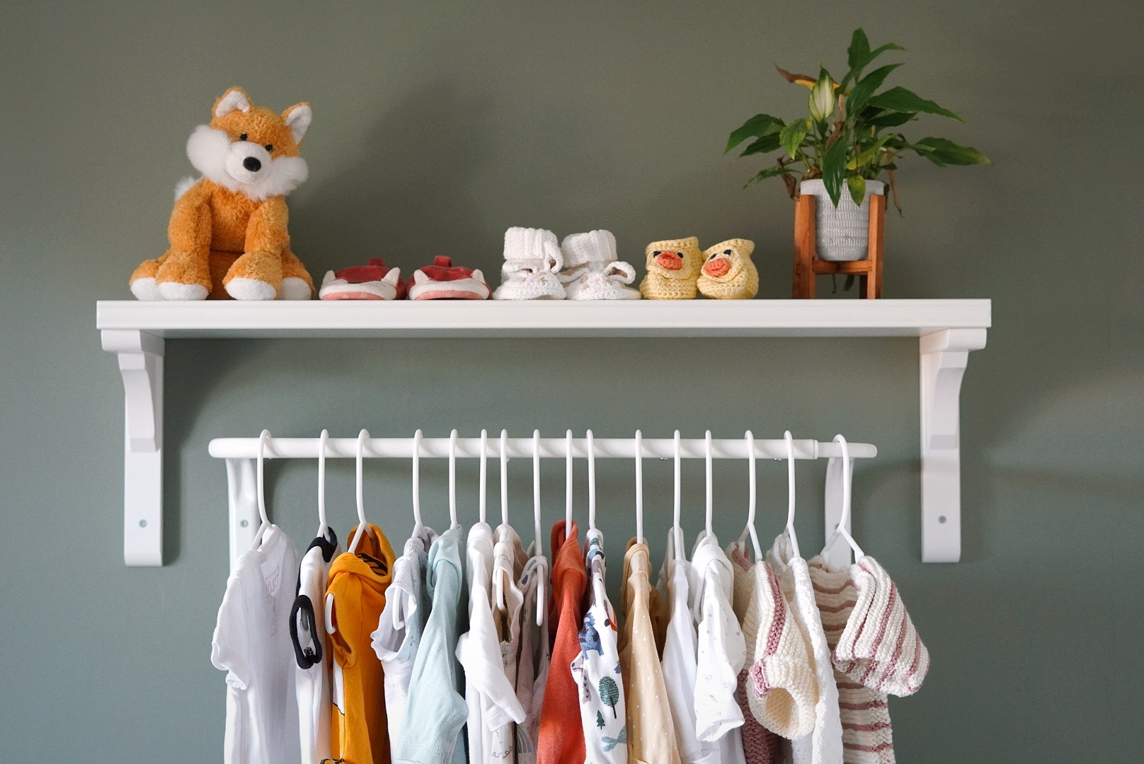 a white shelf above a white clothes rail in a nursery; on the shelf is a toy fox, some knitted booties and a plant. On the clothes rail are some babygrows and knitted baby jumpers