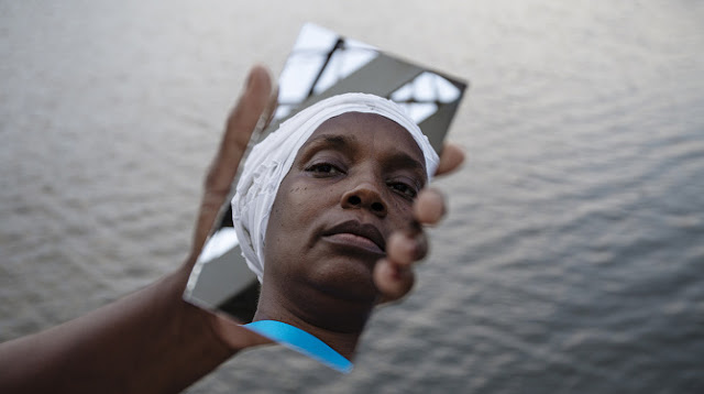 A fotografia contém uma mão segurando um espelho que reflete o rosto de uma mulher negra que está usando um turbante; em seu rosto uma expressão séria. No fundo do cenário há água.