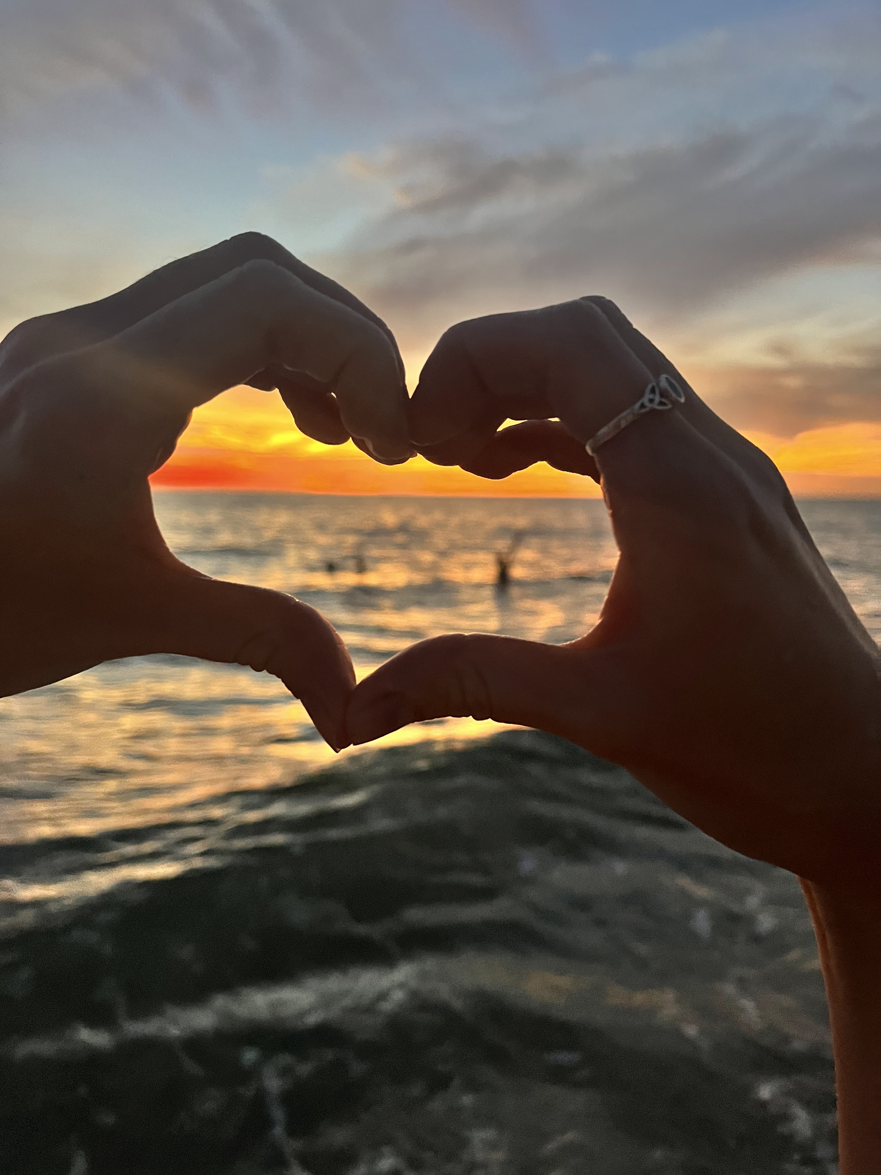 Two hands create a heart. Through them is the ocean, sunset, and two blurry figures.