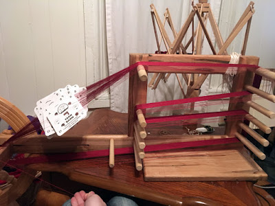 A wooden tablet-weaving loom with a broad band of deep garnet thread wound over some of the pegs and a somewhat disorganized stack of white tablet-weaving cards suspended at left, on a small table with a wooden swift clamped to the back edge.