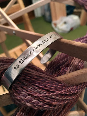 A close-up of a narrow silver cuff bracelet with "to thine own self be true" stamped into the surface in a lowercase sans-serif font, hooked around a small bundle of pink, purple, and white yarn and a stick of a light wooden umbrella swift.