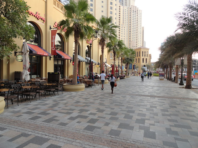 The image shows the walk, JBR. There is a restaurant with outdoor seating, and lots of sky rise buildings in the background. There are also plam trees.