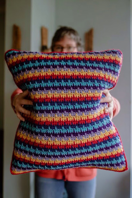 A woman stands holding a large, rainbow coloured, crocheted cushion
