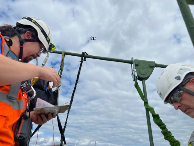 Two transportation workers in hard hats and orange vests navigating a drone high above a cable bridge