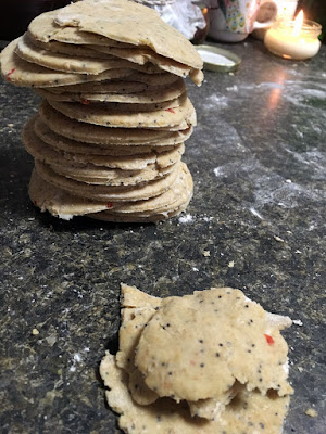 A stack of pale, thin rounds of dough with poppy seeds and red flecks embedded, on a floury dark-green counter with a lit candle in the background and a shorter stack of dough off-cuts in the foreground.