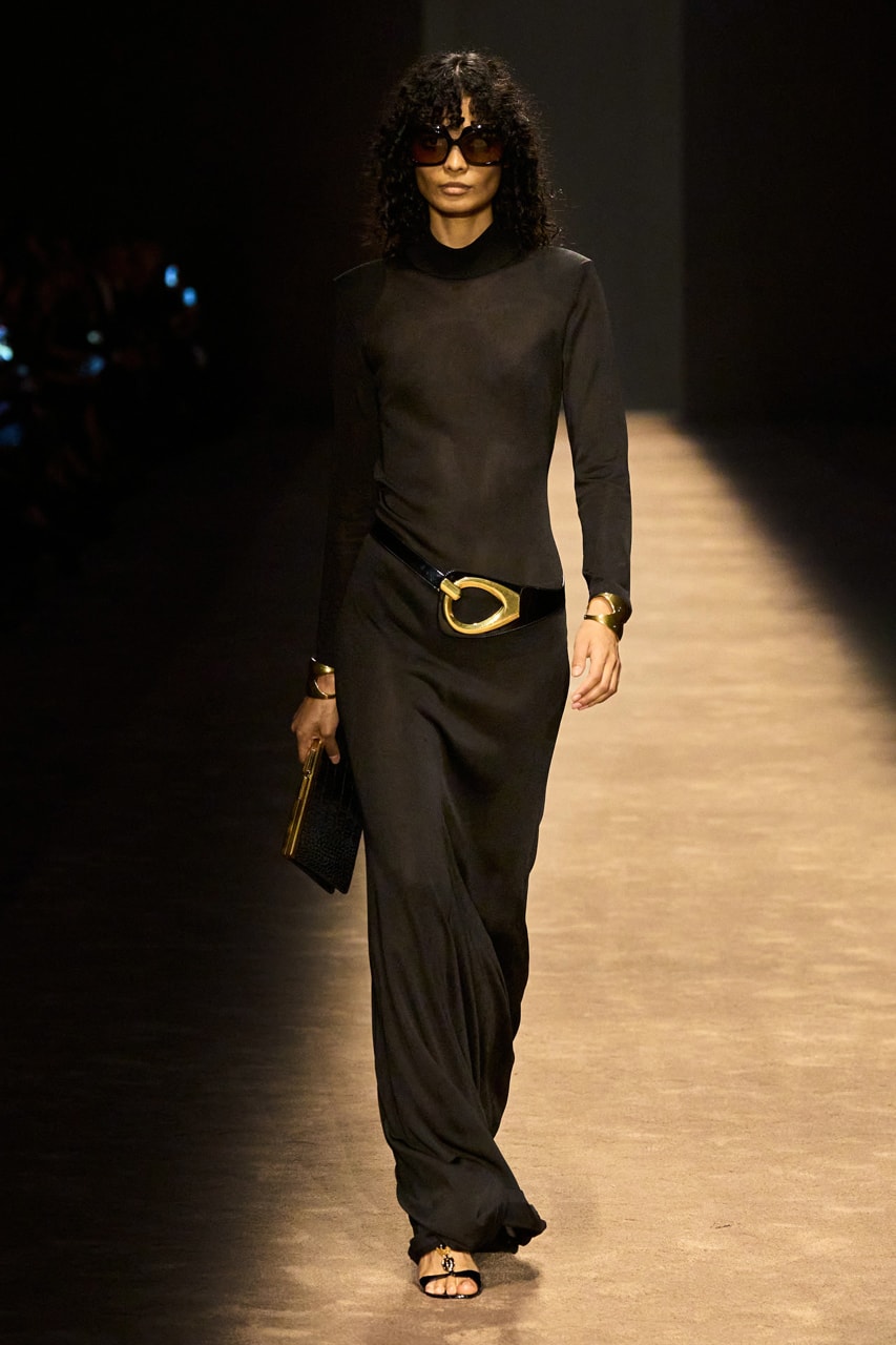 Woman with dark brown curly shoulder length hair with heavy fringe bangs wearing a black long sleeve maxi dress, black strappy sandals, a black leather low-slung belt with gold buckle, and black oversized sunglasses carrying a black clutch on a taupe carpeted runway
