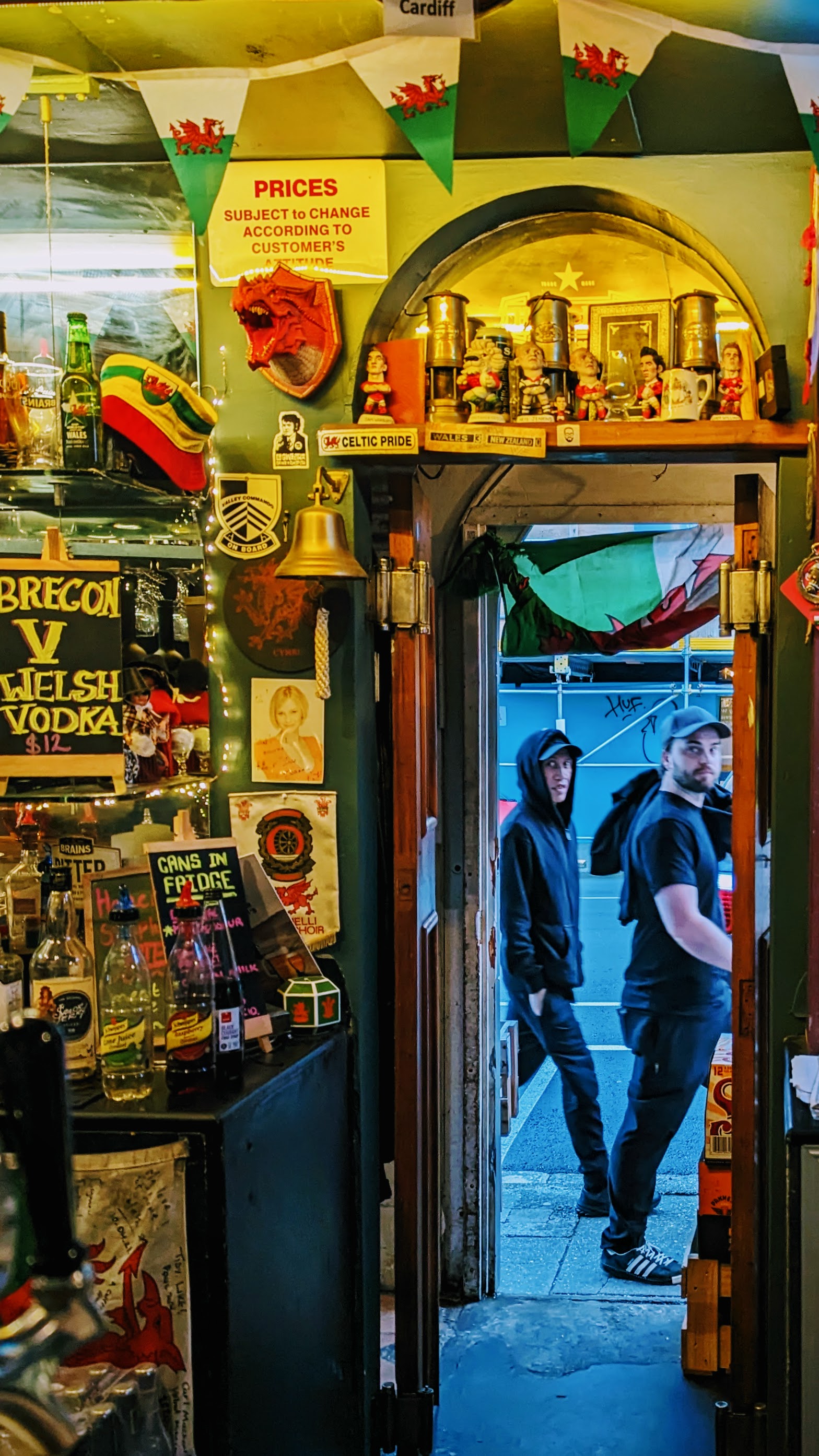 Looking out from a warmly coloured pub through the external bar door to a couple of lads outside staring back in as they walk by. There is a lot of Welsh paraphernalia around the bar.