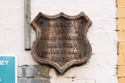 A shield-shaped metal plaque on a wall. It is streaked and darkened, but says 'Borough of Stoke-Newington boundary centre of road 1901'.