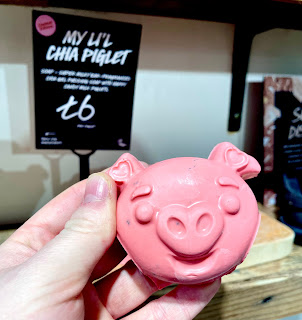 A white hand holding a pink pig shaped soap next to a black rectangular card that says my cute lil chia piglet soap lush in white font on a bright background