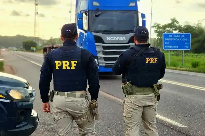 Dois agentes da PRF no acostamento observando caminhão Scania azul passando