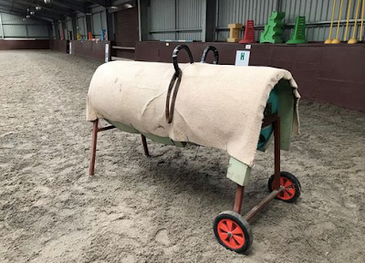 A vaulting barrel in an indoor arena. The barrel is covered in cream carpet. It has two handles and a stirrup leather is threaded through one of these which can be seen because the barrel is slightly side on.