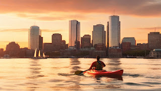 A person kayaking in Boston Harbor at sunset, with the city skyline glowing in the distance. The kayaker wears a life jacket and paddles through the calm water.