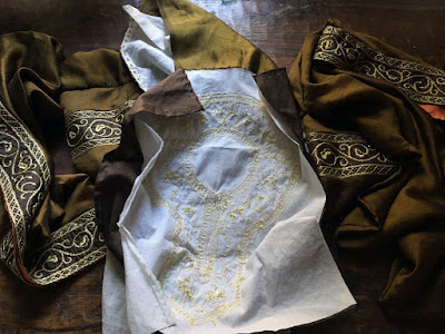 A crumpled mass of green-gold silk with bands of gold floral embroidery on a dark wood table, with a white linen square showing the backside of gold embroidery tracing the shape of a round keyhole neckline, pinned flat in the center of the green-gold fabric.